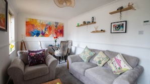 Ein Wohnzimmer mit grauem Sofa, einem kleinen Sessel, einem Couchtisch mit Früchten, einem Gemälde und einem Deckenventilator.