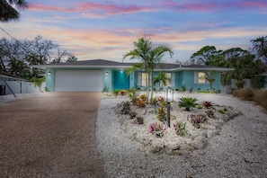 A single-story house with a garage, surrounded by a landscaped garden with various plants and a palm tree. - Mermaid Cove | Backyard Dream: Putting Green, Pool (Siesta Key)