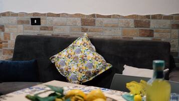 A living room with a sofa, a painting on the wall, a stone wall, a table with a bowl of lemons, and a bottle.