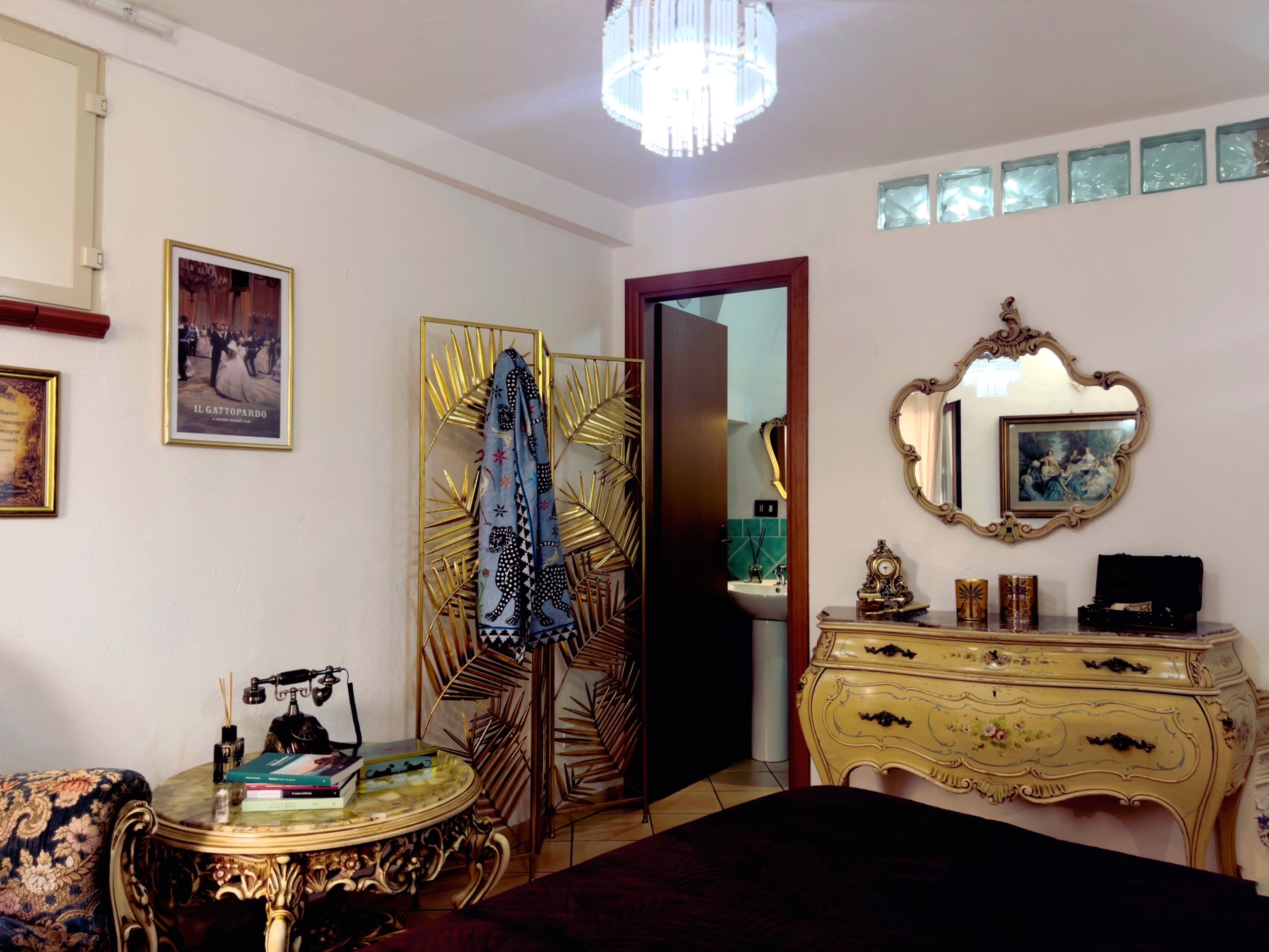 A bedroom with a bed, a vanity with a mirror, a dresser, a round table with books, and a decorative wall panel.