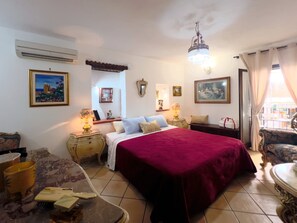 Una habitación de hotel con una cama grande, un candelabro, una ventana con cortinas, una mesita y una silla.