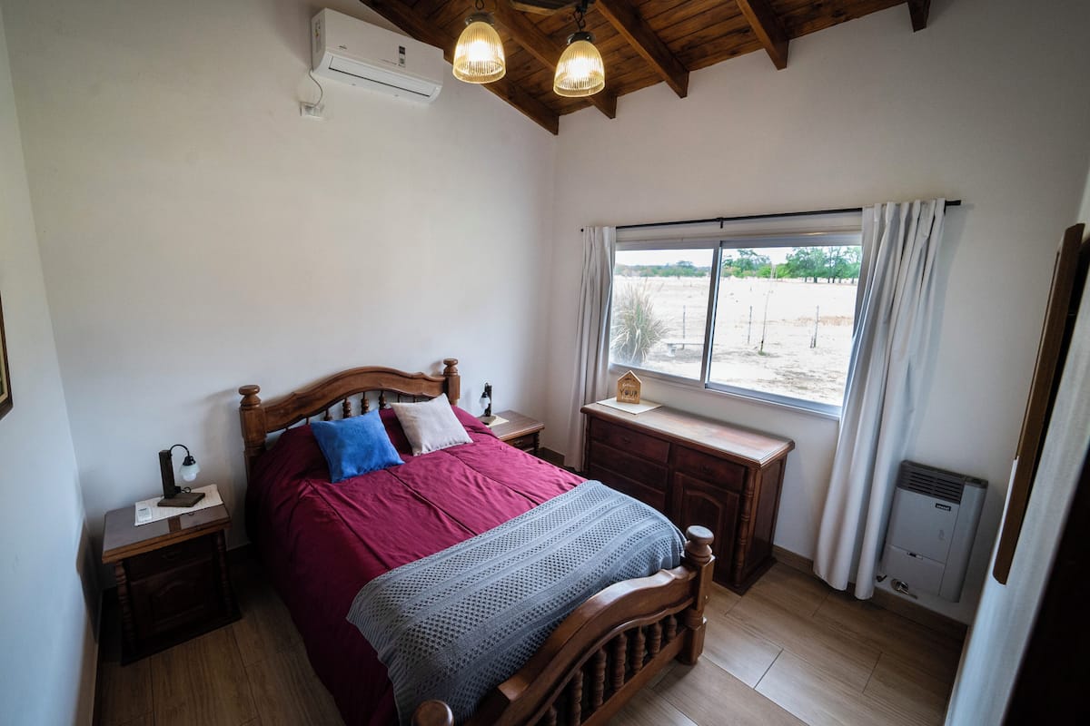 A bedroom with a wooden bed, bedside tables, a window with curtains, an air conditioning unit, and a framed picture on the wall.