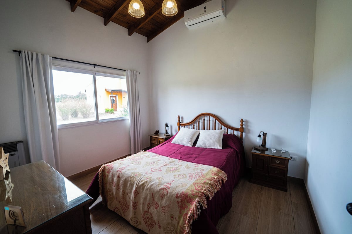 A bedroom with a bed, bedside tables, a desk, a window, and an air conditioning unit.