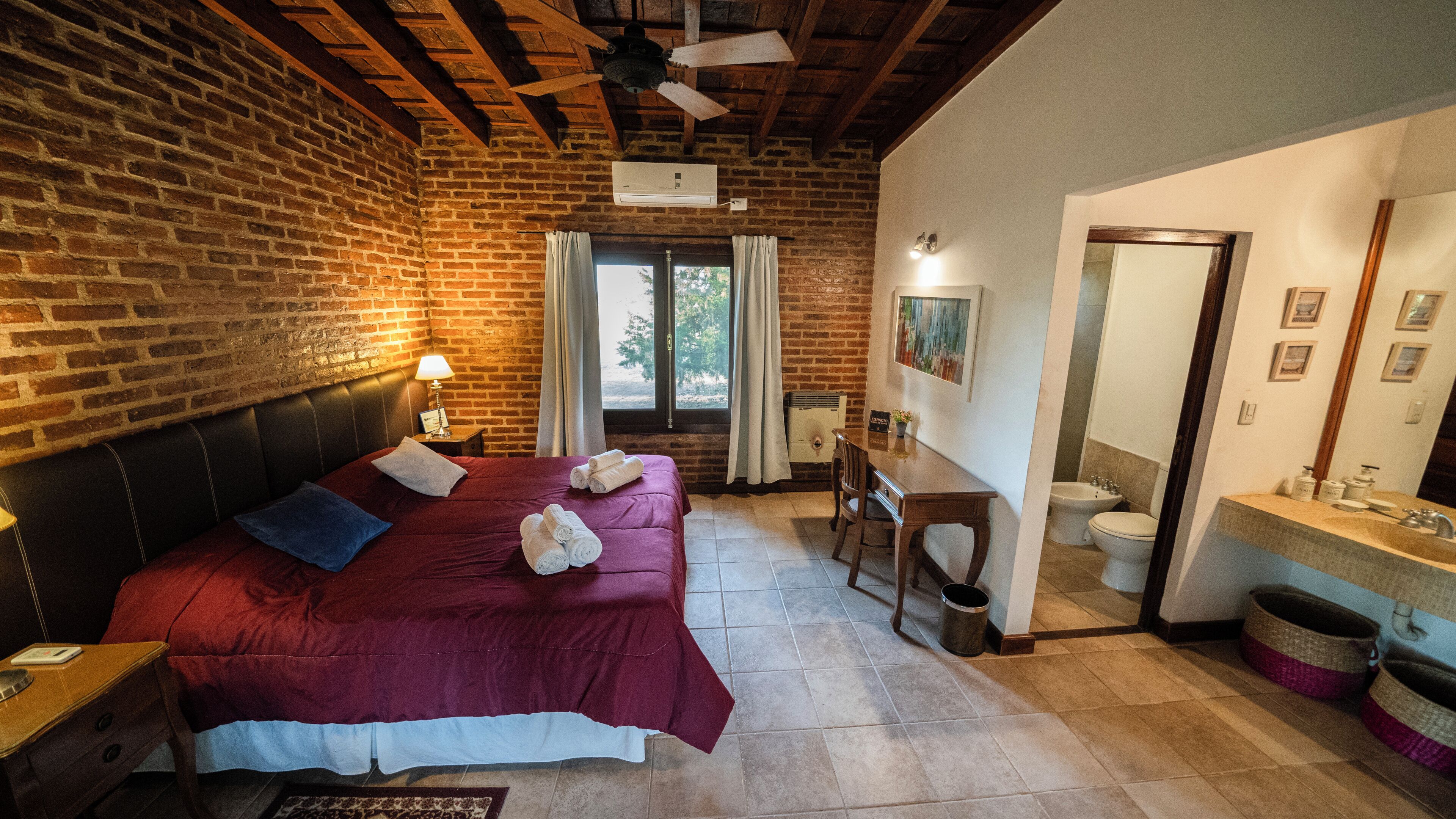 A room with a large bed, a desk, a chair, a bathroom, and a ceiling fan.