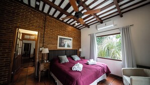 A bedroom with a brick wall, a ceiling fan, a bed with pillows, a bedside table with a lamp, and a window with curtains. - La Soleada Polo Ranch (Mercedes)