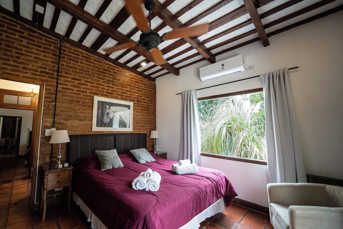 A bedroom with a brick wall, a ceiling fan, a bed with pillows, a bedside table with a lamp, and a window with curtains.