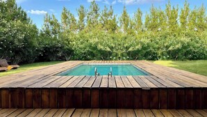 Una piscina rettangolare con un pontile in legno tutt'intorno, sullo sfondo di fitta vegetazione verde e un cielo azzurro limpido.