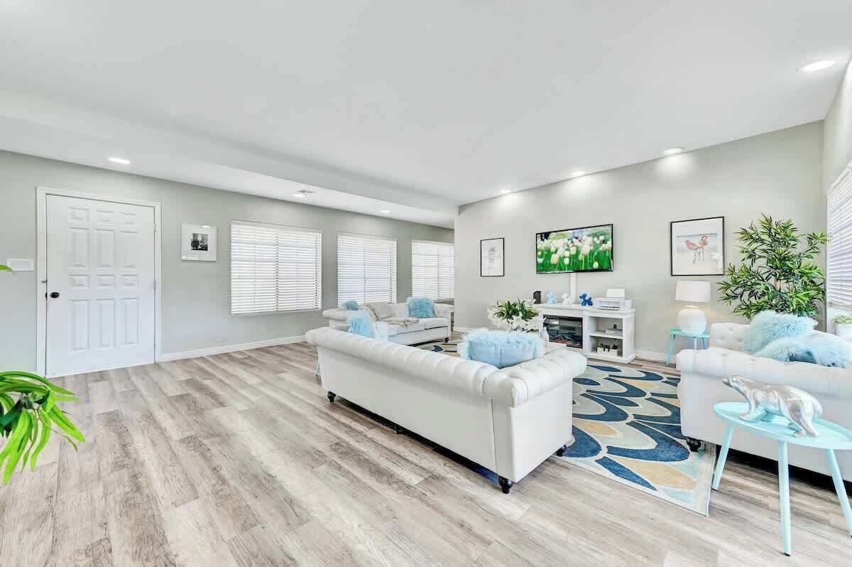A spacious living room with a white sofa, a coffee table, and a TV. There are two windows with blinds, a door, and a plant.