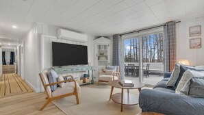 Ein modernes Wohnzimmer mit Flachbildfernseher, einer Couch, einem Couchtisch und einem Essbereich mit Blick auf Bäume.