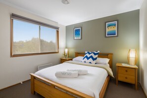 A hotel room with a large bed, a bedside table with a lamp, a window with blinds, and two wall-mounted pictures. - Velora Rye Pet Friendly Home (Rye)