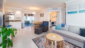 A modern living room with a sofa, ottoman, and round coffee table. There is a dining area with a table and chairs, and a kitchen with white cabinets and a refrigerator. - 204 Jefferson Ave (Cape Canaveral)