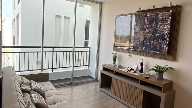 A modern living room with a sofa, a TV mounted on the wall, and a balcony with a cityscape view. - APARTMENT IN PIURA WITH PARK VIEW (Piura)
