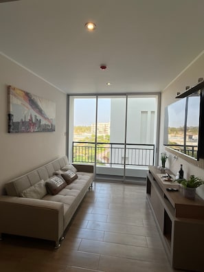 A modern living room with a sofa, a painting, a balcony with a view, and a kitchen area.