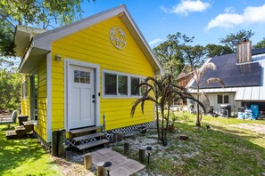 Un bâtiment jaune avec une porte et des fenêtres blanches, une terrasse avec une table et des chaises, et un palmier dans la cour.