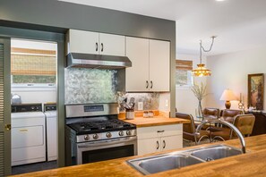 Una cocina compacta con estufa, lavabo y armarios, un comedor con mesa y sillas, y un ventanal con persianas.