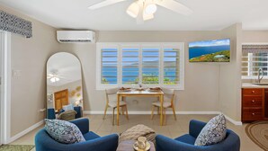 Ein Wohnzimmer mit Deckenventilator, einem Esstisch mit Stühlen, einem großen Fenster mit Jalousien und einer Küchenzeile mit Spüle und Schränken.