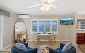Ein Wohnzimmer mit Deckenventilator, einem Esstisch mit Stühlen, einem großen Fenster mit Jalousien und einer Küchenzeile mit Spüle und Schränken.
