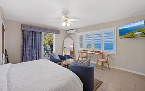 Ein Schlafzimmer mit Bett, Deckenventilator, Fenster mit Rollläden, Esstisch mit Stühlen und Meerblick.