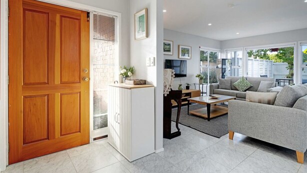 A modern living room with a grey sofa, a wooden door, and a white wall with a window. - Remuera 3BR Elegance Home with Outdoor Living (Auckland Central Suburbs)