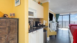 Cocina compacta con armarios blancos, microondas y lavamanos. Tiene un balcón con una cama roja y vista al mar.