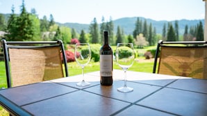 Ein Tisch mit einer Weinflasche und zwei Gläsern auf einer gefliesten Terrasse mit Stühlen, mit Blick auf einen Garten und Berge.