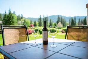 Ein Tisch mit einer Weinflasche und zwei Gläsern auf einer gefliesten Terrasse mit Stühlen, mit Blick auf einen Garten und Berge.