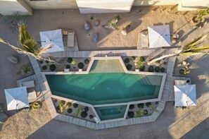 Vista aérea de un área de piscina con una pileta rectangular, rodeada por un deck de madera, palmeras y sombrillas blancas.