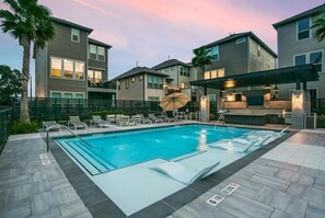 A swimming pool with lounge chairs, a covered patio area, and a view of multi-story buildings.