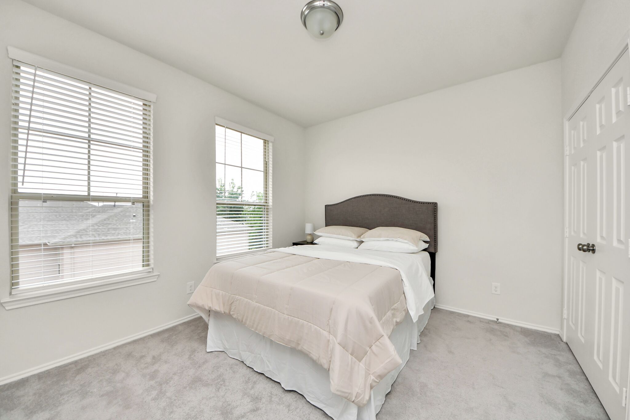 A bedroom with a bed, a nightstand, a window with blinds, and a door.
