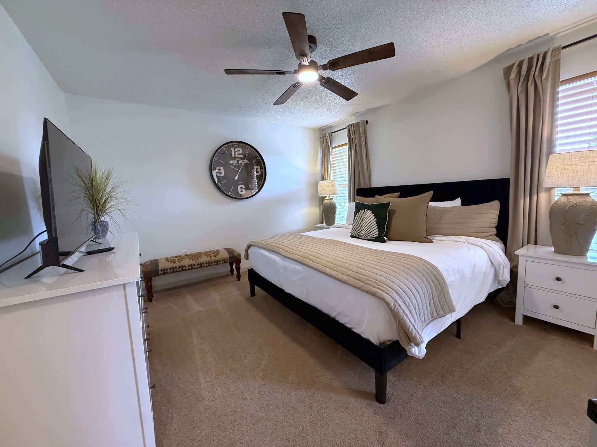 A bedroom with a bed, a ceiling fan, a clock, a television, a bedside table with a lamp, and a window with curtains.