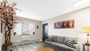 A living room with a grey sofa, a round table, a potted plant, a radiator, and a painting on the wall.