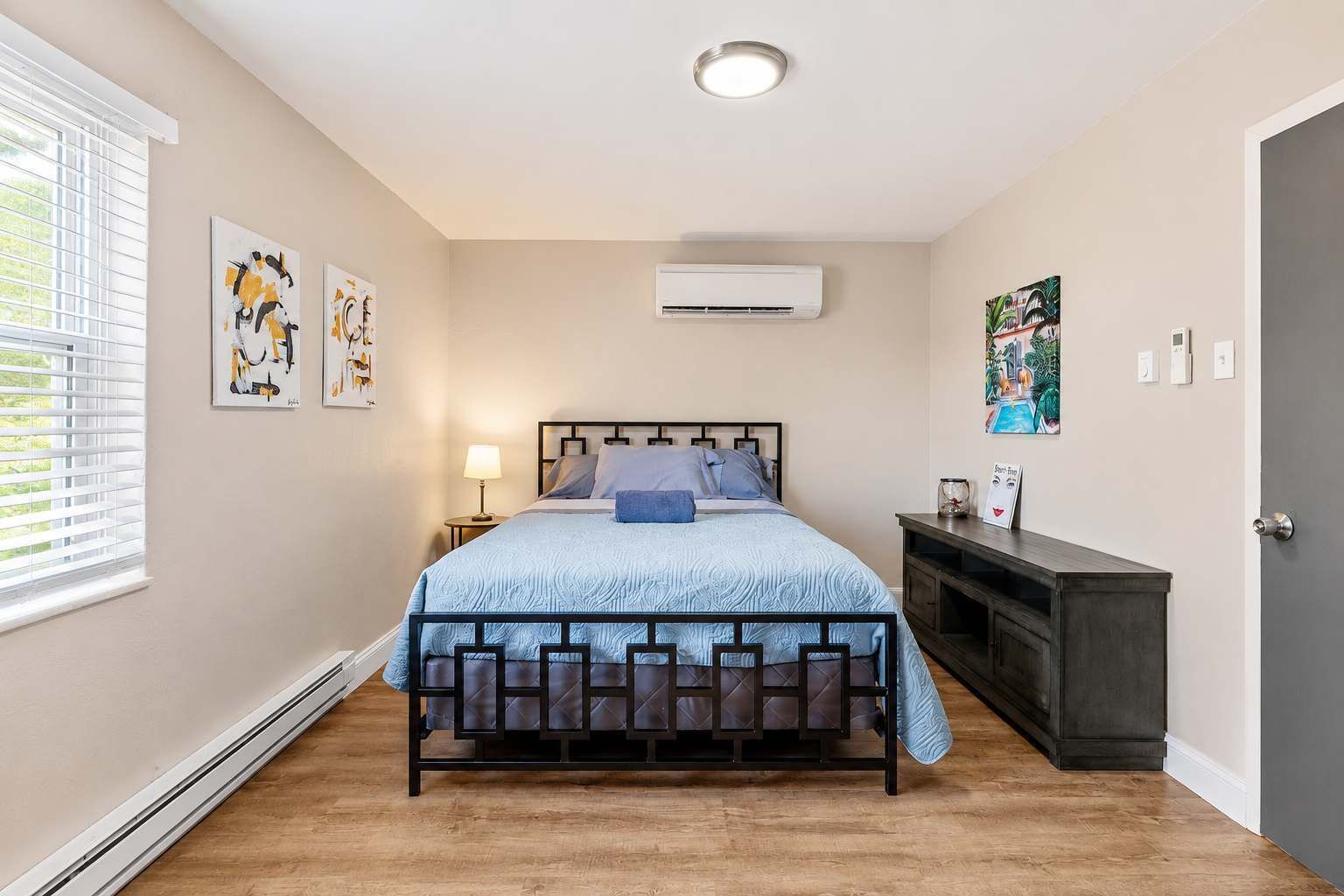Een slaapkamer met een bed, nachtkastje, lamp, kunstwerk, airconditioning en een donker houten dressoir.