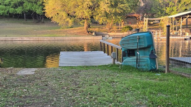 A calm lake with a dock, a green boat, and a fenced area with trees. - Hidden Cove RV Site On Lake LBJ. (Kingsland)