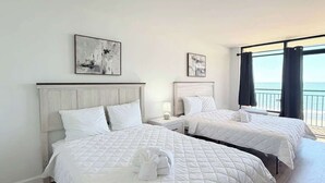 Ein Hotelzimmer mit zwei Betten, weißer Bettwäsche, Kunstwerken an den Wänden, einem Nachttisch mit Lampe und Meerblick durch große Fenster.
