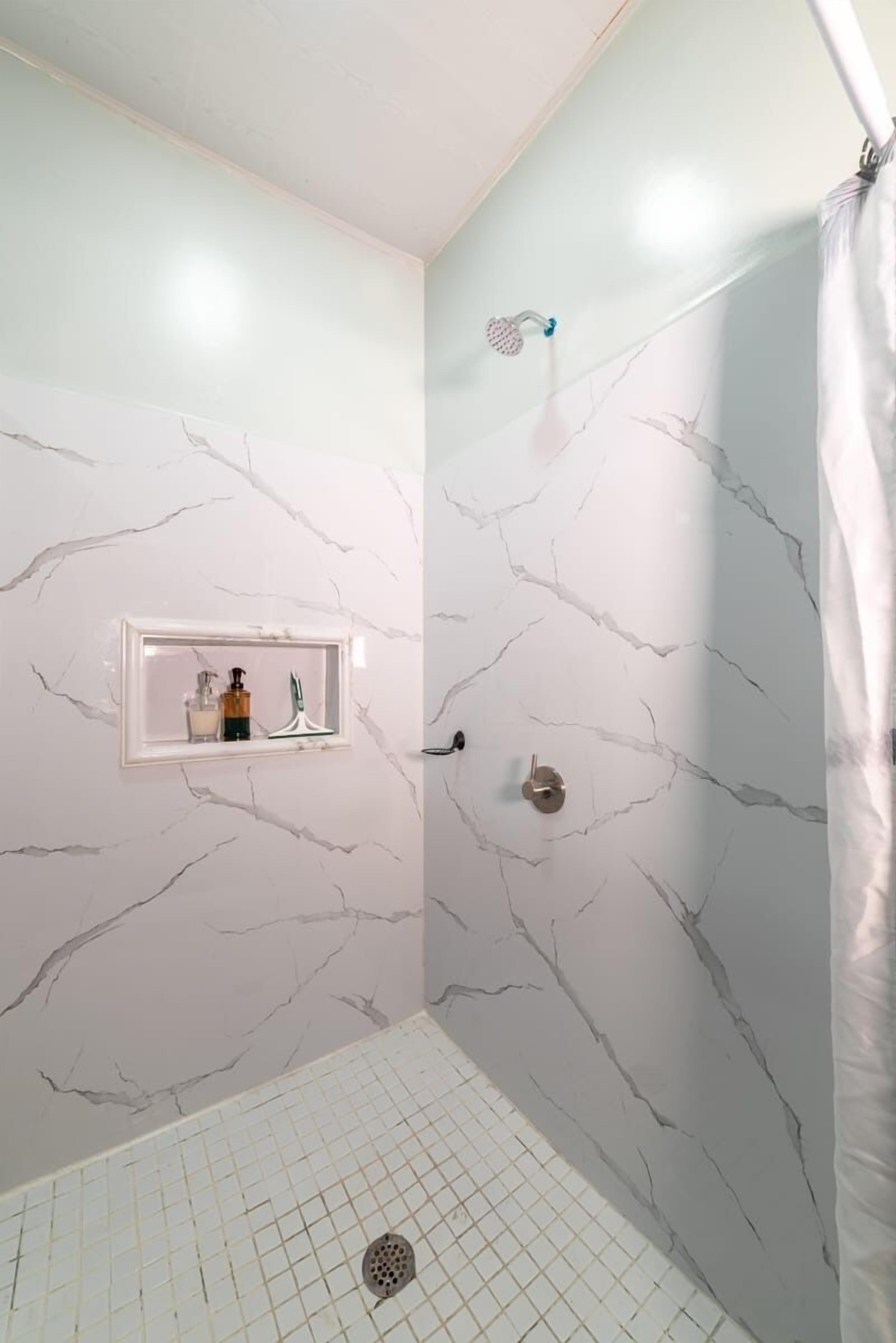 A bathroom with white marble walls, a shower with a curtain, and a shelf with toiletries.