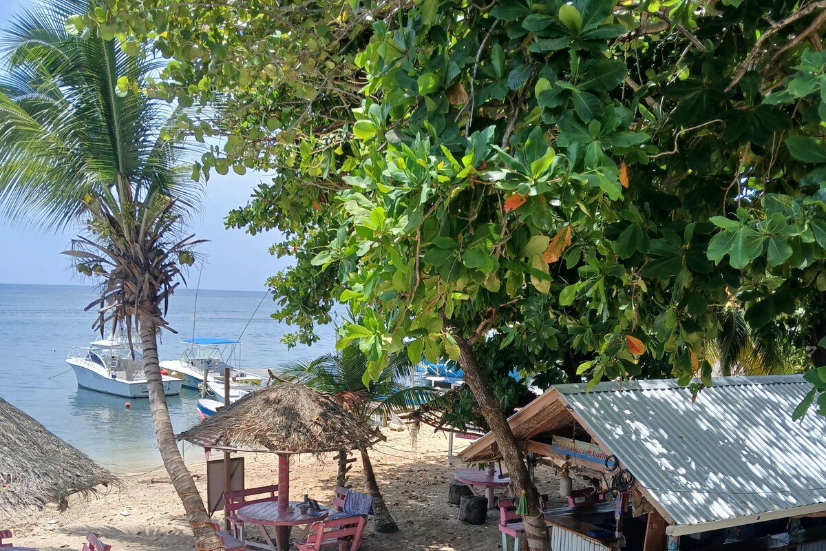 Ein Strandabschnitt mit einer strohgedeckten Hütte, einer kleinen Bar und einer Sitzgelegenheit mit Tischen und Stühlen.