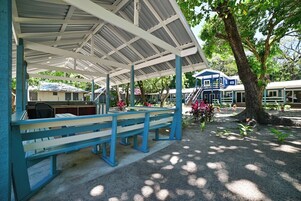 A covered outdoor seating area with benches, a building with a blue facade, and a tree in the background.