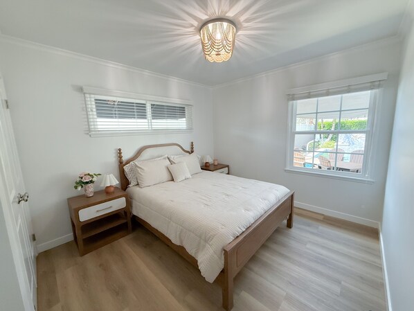 A bedroom with a wooden bed, bedside tables, a nightstand, a chandelier, a window with blinds, and a view of a pool outside.