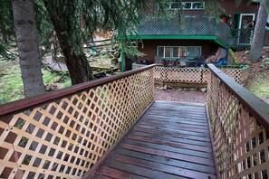 Um deck de madeira leva a uma cabana com telhado verde, cercada por árvores.