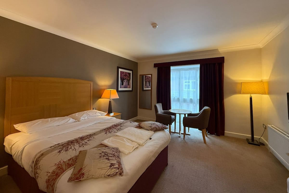 Habitación de hotel con una cama grande, dos lámparas de noche, una mesita con sillas y una ventana con cortinas.