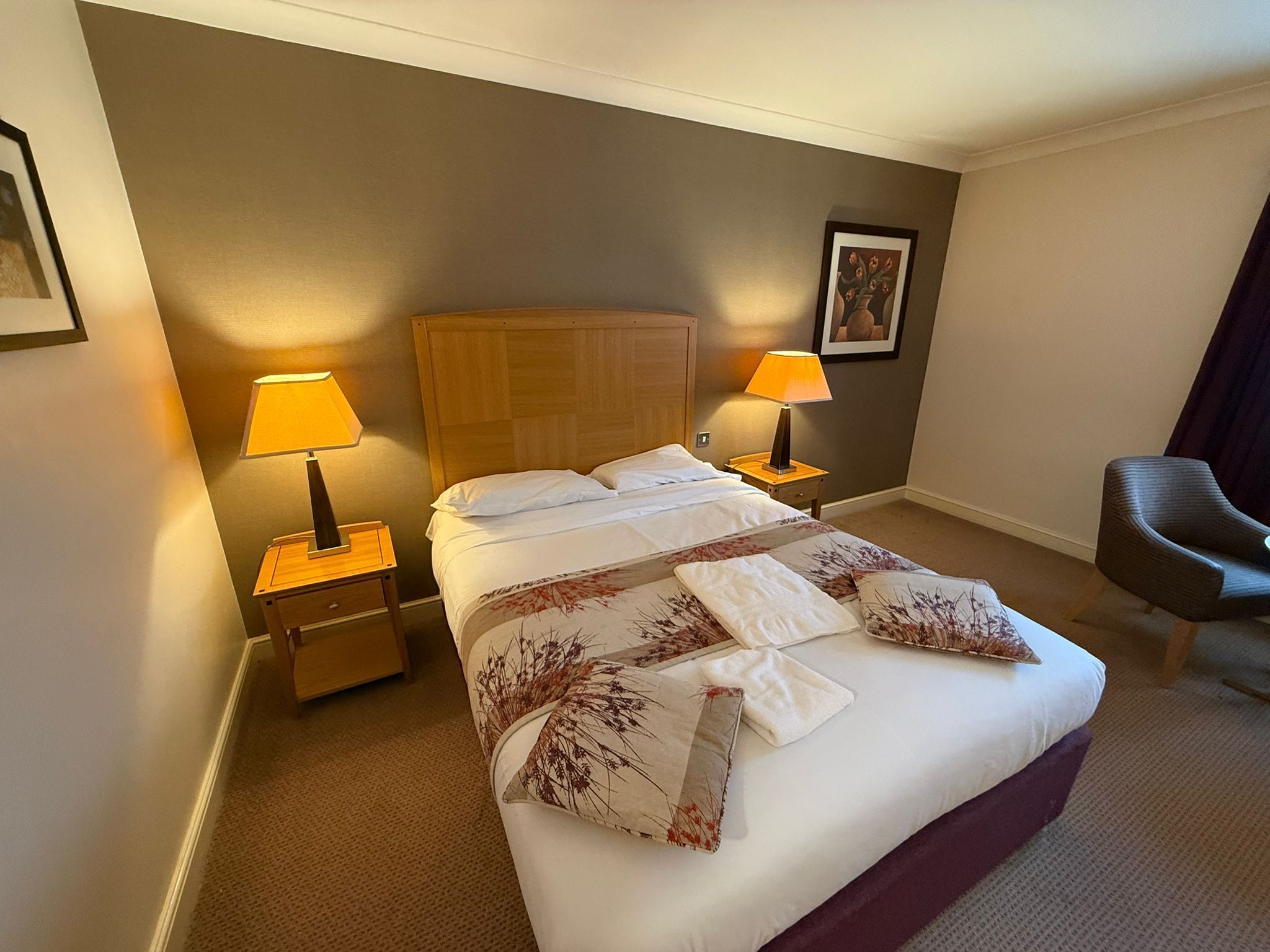 A hotel room with a large bed, two bedside lamps, a wooden headboard, a chair, and a framed picture on the wall.