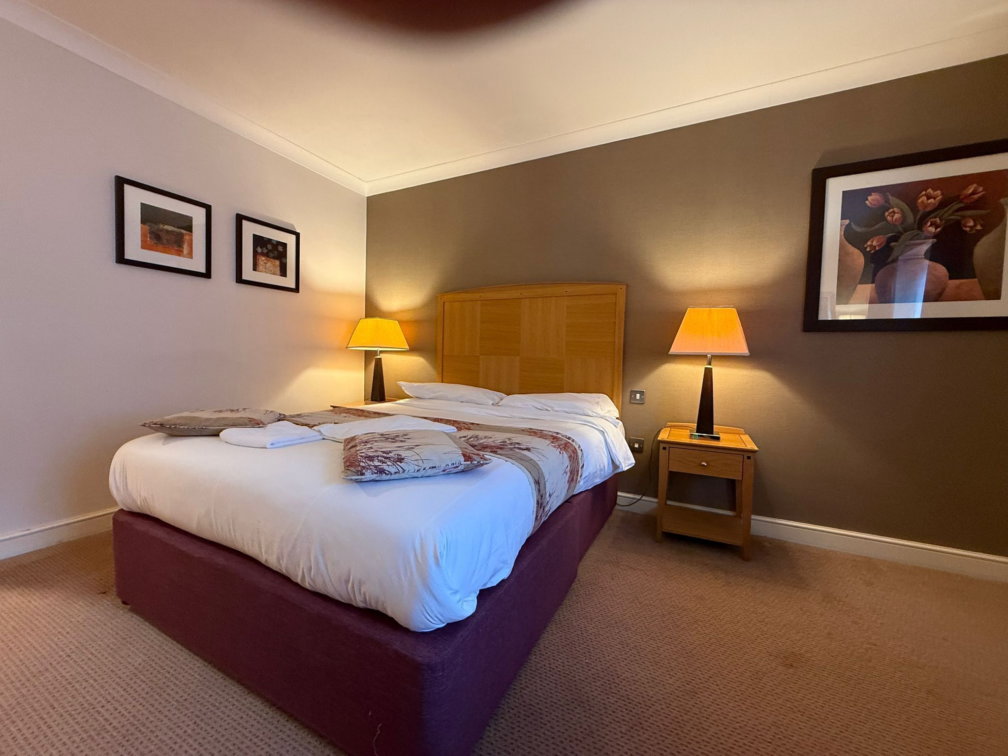 A hotel room with a bed, two bedside lamps, a wooden headboard, a carpeted floor, and two framed pictures on the wall.