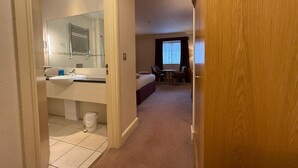 A hotel room with a bathroom, a bed, a desk, and a window.