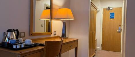 A hotel room with a wooden desk, a chair, a lamp, a mirror, and a door with a fire alarm sign.