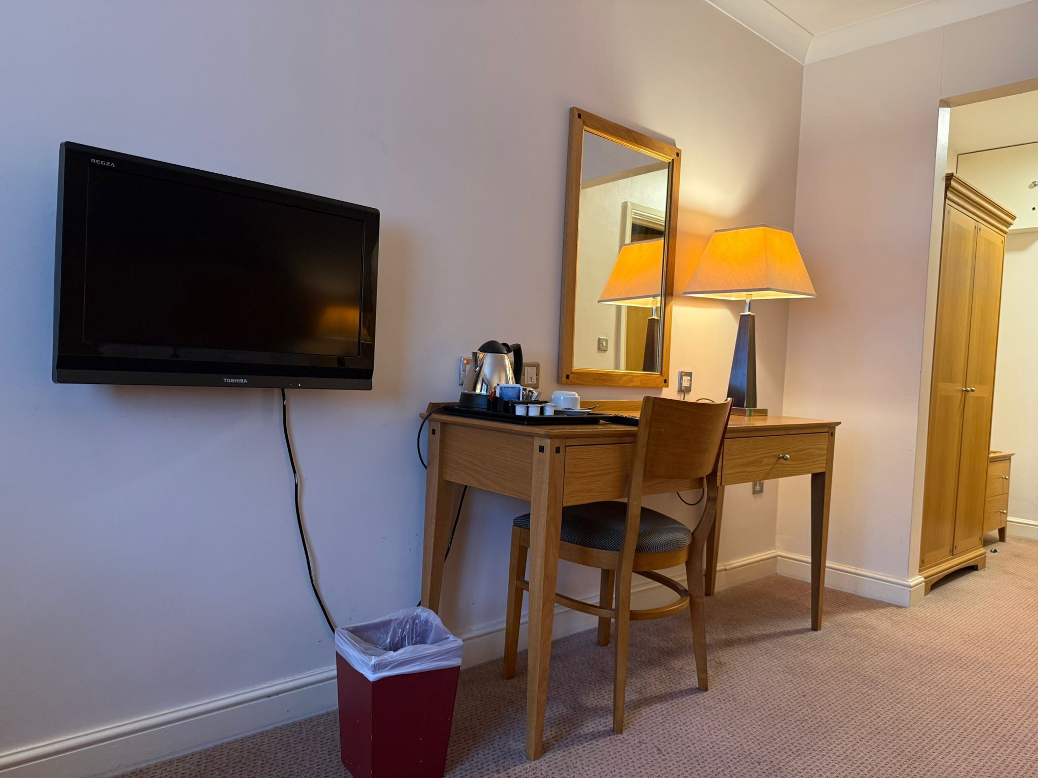 A hotel room with a desk, chair, TV, lamp, and wardrobe.