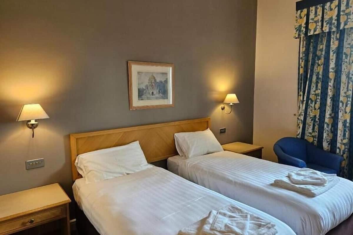 Habitación de hotel con dos camas, una mesita de noche, una lámpara, un cuadro enmarcado en la pared y una ventana con cortinas.