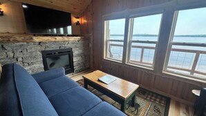 A cozy room with a stone fireplace, a flat-screen TV, a blue sofa, and large windows overlooking a body of water. - Cabin on the beach with lakeviews and fireplace (AU TRAIN)