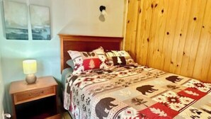 A bedroom with a wooden headboard, a bedside table with a lamp, and a patterned bedspread. - Cabin on the beach with lakeviews and fireplace (AU TRAIN)