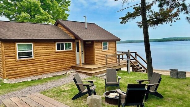 A log cabin with a deck, outdoor seating, and a view of a lake. - Cabin on the beach with lakeviews and fireplace (AU TRAIN)