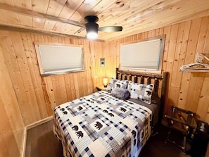 Una stanza con rivestimento in legno, un letto, due finestre con persiane, una ventola a soffitto e un appendiabiti.